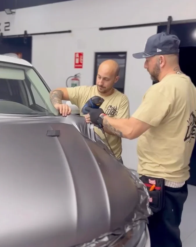 Dos técnicos aplicando vinilo sobre el capó de un vehículo gris con ayuda de calor