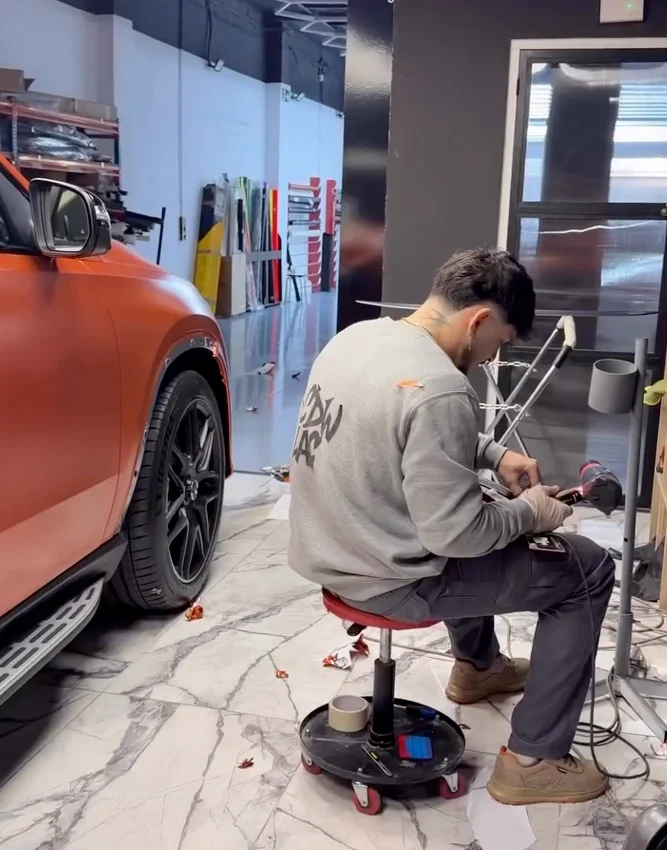Técnico preparando herramientas y material de vinilo junto a un SUV Mercedes naranja satinado